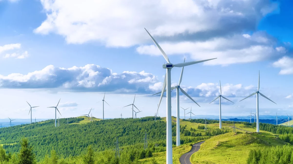 Wind turbines on a lush green hillside under a bright blue sky with scattered white clouds. A winding road leads through the landscape, showcasing a sustainable energy scene.